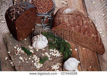 Close up photo of rustic bread sprinkled with sunflower seeds on sackcloth, garlic, dill, oat flakes and jar with buckwheat on planked wooden table