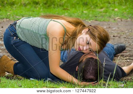 Young man lying down with medical emergency, woman sitting by his side checking for breath, outdoors environment.