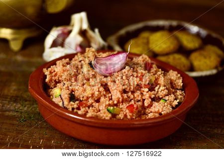closeup of an earthenware casserole whit couscous with vegetables and some falafel in a plate in the background, on a wooden table