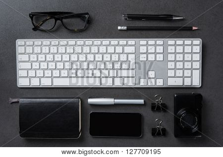 High angle view of a professional desktop. Top view of tidy desk in a business office. Close up of keyboard, organizer, camera, pen, smartphone, pencil and glasses on blackboard background.