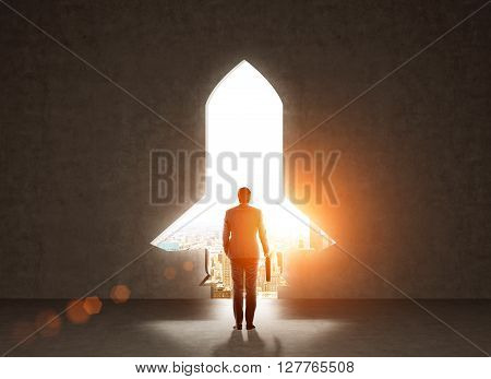 Start up concept with businessman holding briefcase and standing in front of rocket shaped gap in wall revealing sunlit New York city view
