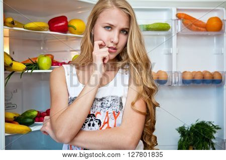 Beautiful young girl standing at the open refrigerator with fruits and vegetables and thinking