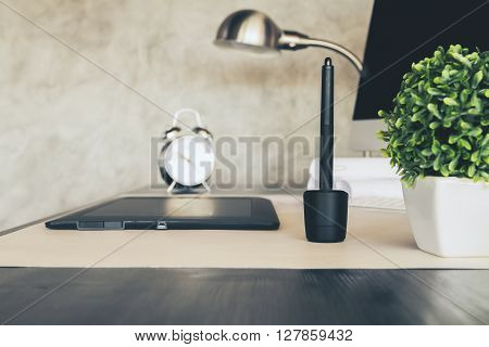 Sideview of desktop with graphic tablet plant alarm clock lamp and computer. Concrete wall background