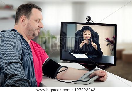 Virtual therapist carefully watches blood pressure figures of her patient. She wears headset because works online. Middle-aged man uses blood pressure gauge for measurement in front of monitor with doctor. Horizontal indoors shot