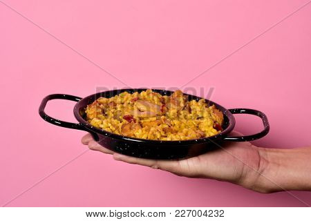 closeup of a young caucasian man holding a typical spanish paella valenciana in a typical paella pan, against a pink background with some blank space on top