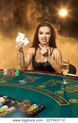 Gorgeous Emotional Young Woman In A Black Dress Sitting At Poker Table With Glass Of Champagne. Let 