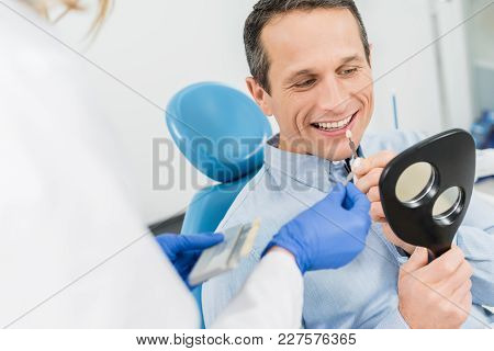 Male Patient Choosing Tooth Implant Looking At Mirror In Modern Dental Clinic