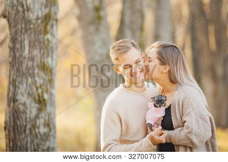 Autumn Photo, Couple In Love Hugs And Holds Dwarf Dog On Background Of Yellow Leaves, Girl Bites Man