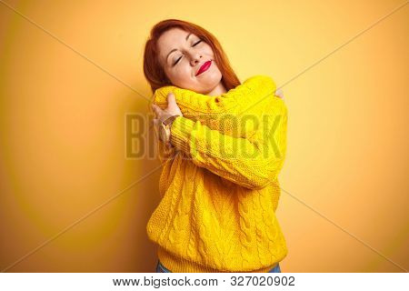 Beautiful redhead woman wearing winter sweater standing over isolated yellow background Hugging oneself happy and positive, smiling confident. Self love and self care