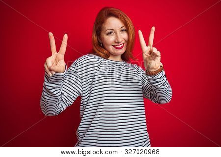 Young redhead woman wearing strapes navy shirt standing over red isolated background smiling looking to the camera showing fingers doing victory sign. Number two.