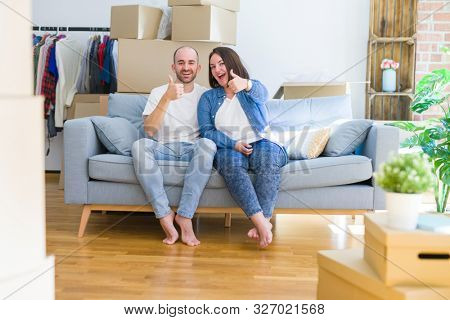 Young couple sitting on the sofa arround cardboard boxes moving to a new house doing happy thumbs up gesture with hand. Approving expression looking at the camera showing success.