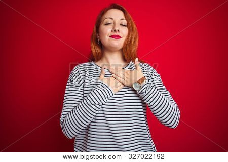 Young redhead woman wearing strapes navy shirt standing over red isolated background smiling with hands on chest with closed eyes and grateful gesture on face. Health concept.