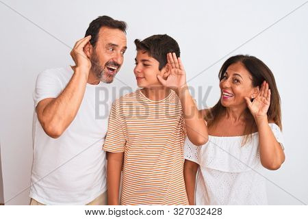 Family of three, mother, father and son standing over white isolated background smiling with hand over ear listening an hearing to rumor or gossip. Deafness concept.