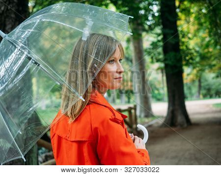 Caucasian Girl With Red Gabardine Walks Through A Park Under A Transparent Umbrella.