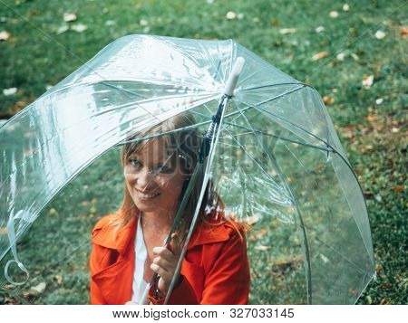 Caucasian Girl With Red Gabardine Walks Through A Park Under A Transparent Umbrella.