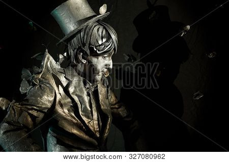 Side View Of Professional Pantomimist In Painted Suit And Hat, Standing Still With Black Wall Behind