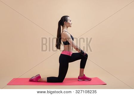 A Young Attractive Girl With A Ponytail, A Brunette, Stands On One Knee, Doing Stretching On A Pink 