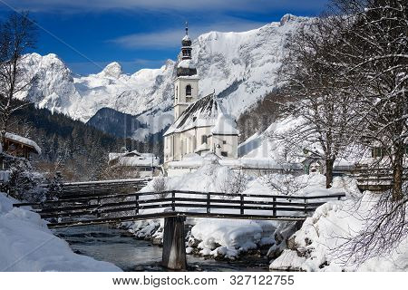 St. Sebastian Church Next To A Mountain Stream With A Bridge And Reiter Alps Mountains In The Backgr