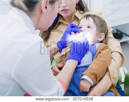 The Doctor Looks At The Teeth Of A Small Child. The Baby Is Sitting In The Arms Of Mom. Dentist Exam