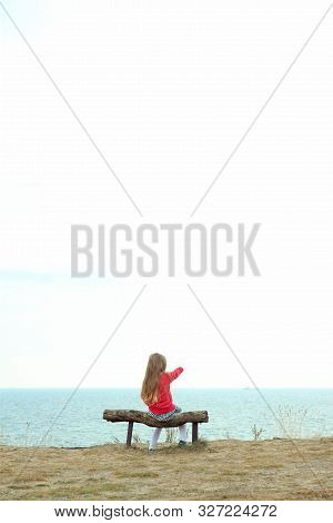 A Little Girl Sits Alone. Solo Travelling. A Tourist Sits On A Wooden Bench On A Cliff Above The Sea