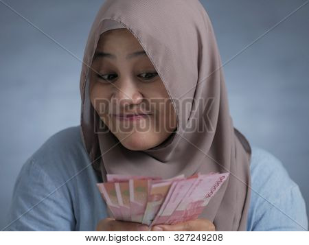 Portrait Of Indonesian Muslim Woman Holding Rupiah Money, Smiling And Thinking Gesture