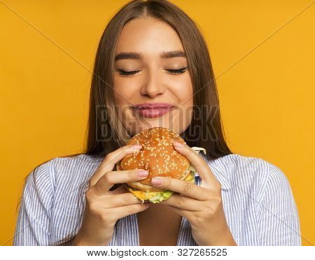 Cheat Meal. Happy Girl Eating Burger Standing Over Yellow Background. Studio Shot