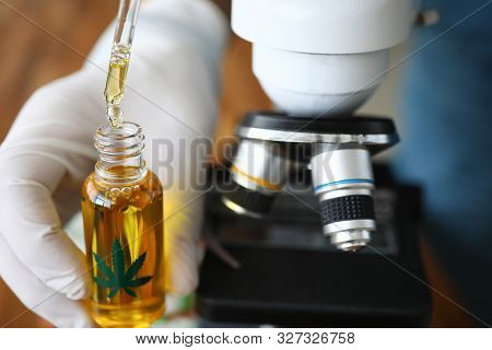 Focus On Male Hands In Gloves Holding Flask With Cannabinoid Oil And Dripping Drops In Glass Bottle.