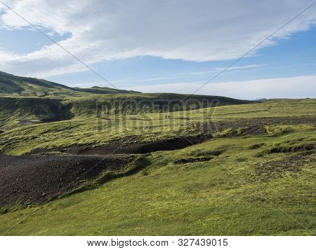 Two Hikers Backpack Travelers In Volcanic Landscape With Green Hills, Meadow And Lava Gravel Ground 
