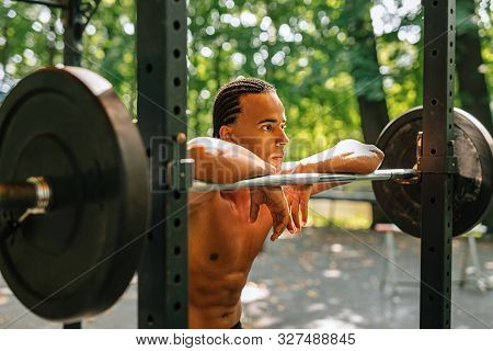 Side View Of A Bare-chest Athlete Standing Outdoors Leaning On A Weight Bar