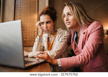 Image of a shocked emotional two business women colleagues indoors in cafe using laptop computer.