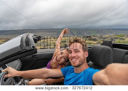 Car couple taking selfie photo with phone on road trip driving cabriolet sports car on summer vacation with arms up in fun. Asian girl, Caucasian man dancing in open roof car.