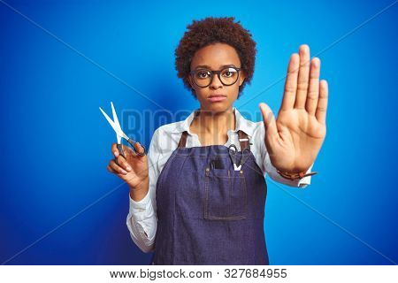 African american hairdresser woman holding scissors over blue isolated background with open hand doing stop sign with serious and confident expression, defense gesture