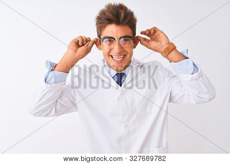 Young handsome sciencist man wearing glasses and coat over isolated white background Smiling pulling ears with fingers, funny gesture. Audition problem