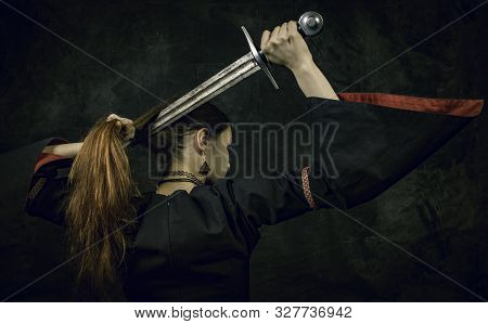Portrait Of Young Long Haired Woman Wearing A Medieval Costume. Standing Pose With A Sword At Her Ha