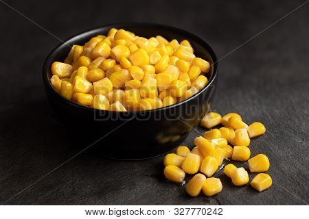 Canned Sweet Corn In A Black Ceramic Bowl Isolated On Black Slate Next To Spilled Sweet Corn.