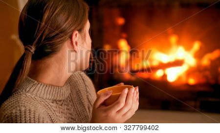 Portrait Of Woman In Woolen Sweater Holding Cup Of Hot Tea And Looking At Burning Firepalce