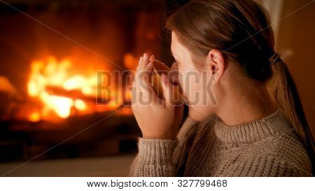 Portrait Of Young Woman In Sweater Dirnking Tea And Enjoying Burning Fire In Fireplace At Home