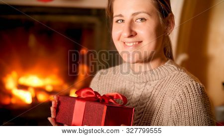 Portrait Of Smiling Happy Woman Sitting By The Firepalce And Holding Red Gift Box