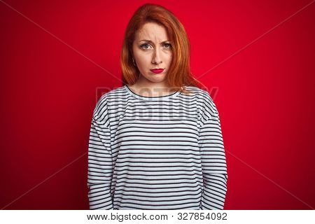 Young redhead woman wearing strapes navy shirt standing over red isolated background skeptic and nervous, frowning upset because of problem. Negative person.