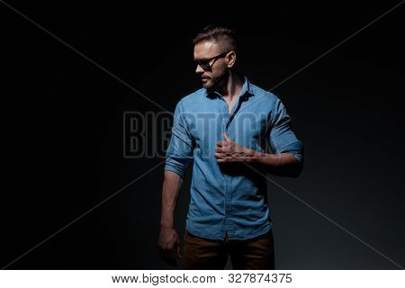 side view of a cool casual man wearing blue shirt standing and fixing shirt while looking down with a dramatic look against black studio background