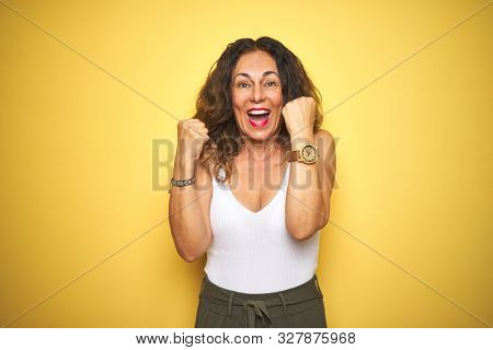 Middle age senior woman showing wrist watch over yellow isolated background screaming proud and celebrating victory and success very excited, cheering emotion