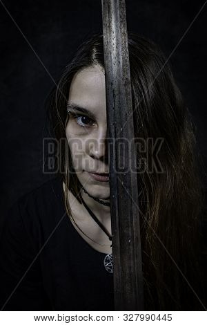 Portrait Of Young Long Haired Woman In Standing Pose Holding A Sword Over Her Left Eye At Dark Studi