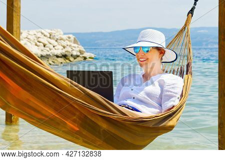 Work From Anywhere. Young Smilyng Woman, Female Freelancer In Straw Hat Working On Laptop On Beach.
