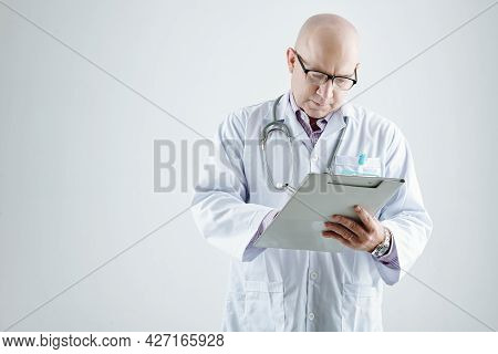 Mature Doctor In White Coat Making Notes In Medicl Card Standing Against The White Background