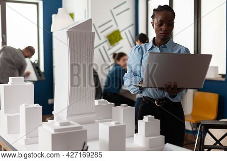 African American Architect At Business Workplace Looking At Laptop And Blueprint Plan For Building M
