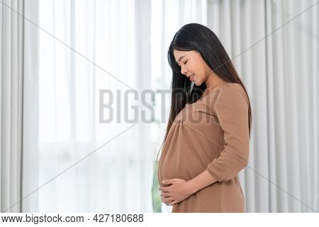 Asian Happy Pregnant Woman Is Standing Beside Windows And Touching Her Belly. Pregnancy, Motherhood,