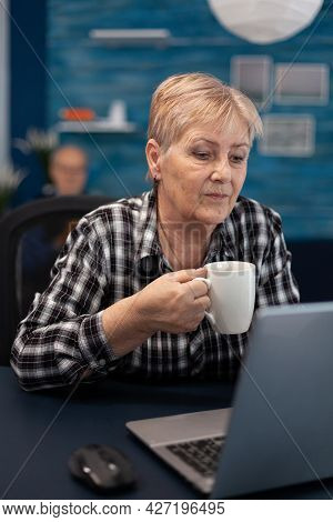 Senior Entrepreneur Woman Reading On Computer Enjoying A Cup Of Coffee And Husband And Reading A Boo