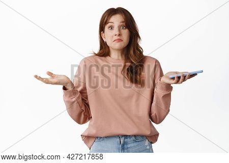 Portrait Of Confused Redhead Woman Shrugging Shoulders, Holding Mobile Phone And Look Clueless, Cant