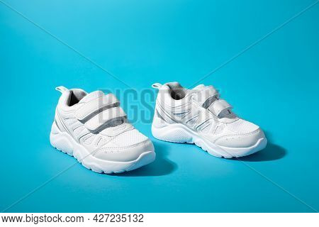 Side View Of Two White Unisex Sneakers With Velcro Fasteners For Quick Shoeing On A Blue Paper Backg