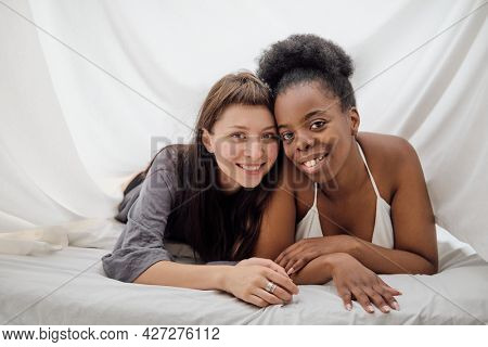 Two cheerful lesbian girls lying close to each other in bed in front of camera